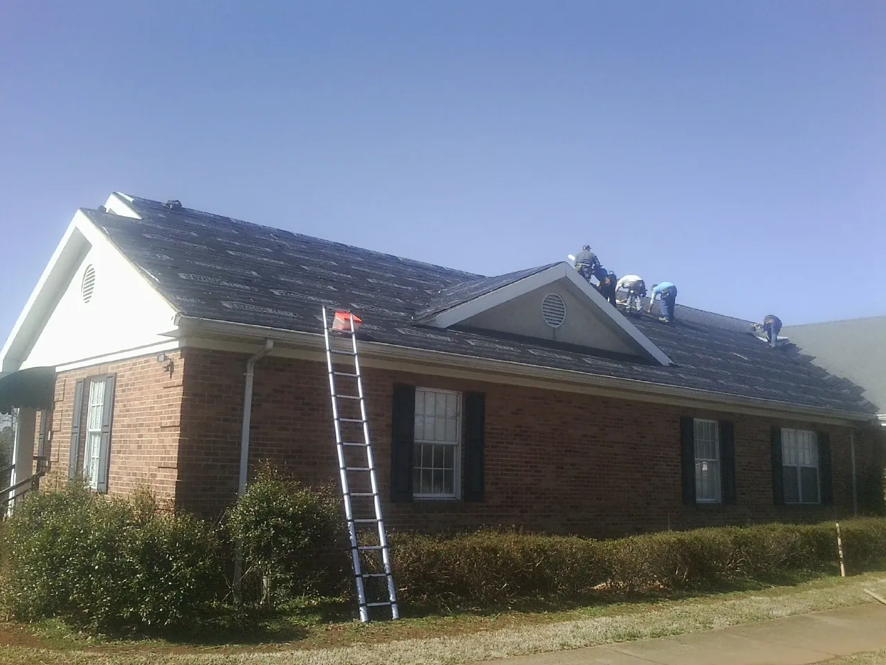 Crew tearing shingles off a brick ranch roof with a ladder set — Metal Roof Repair work by our crew in Delton