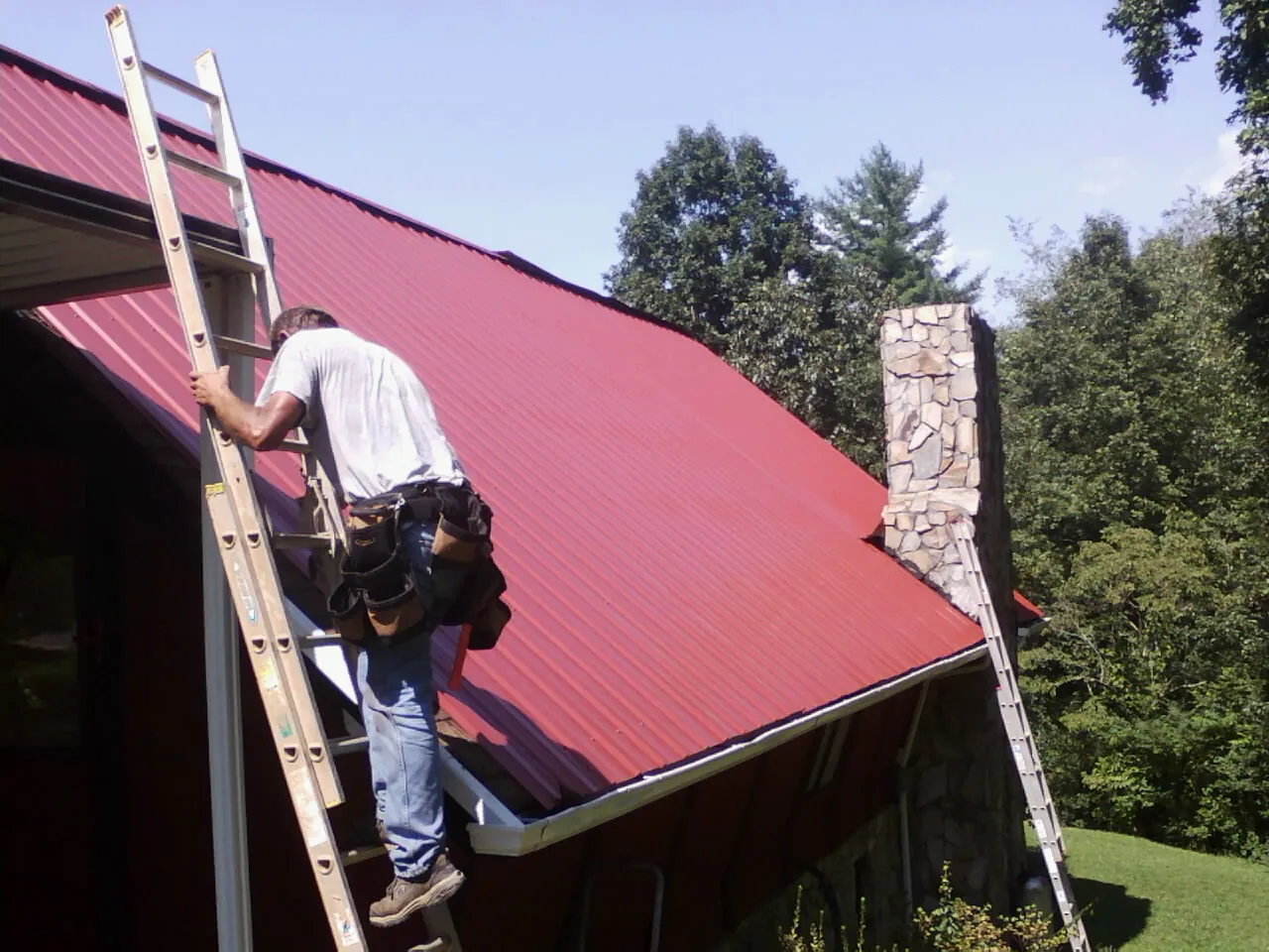 Roofer on a ladder beside a red metal roof and stone chimney — Soffit & Fascia Repair by Roofing Brothers in Delton
