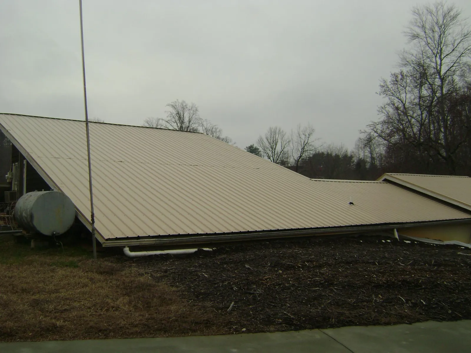 Wide tan metal roof plane seen from the ground beside a propane tank in Delton